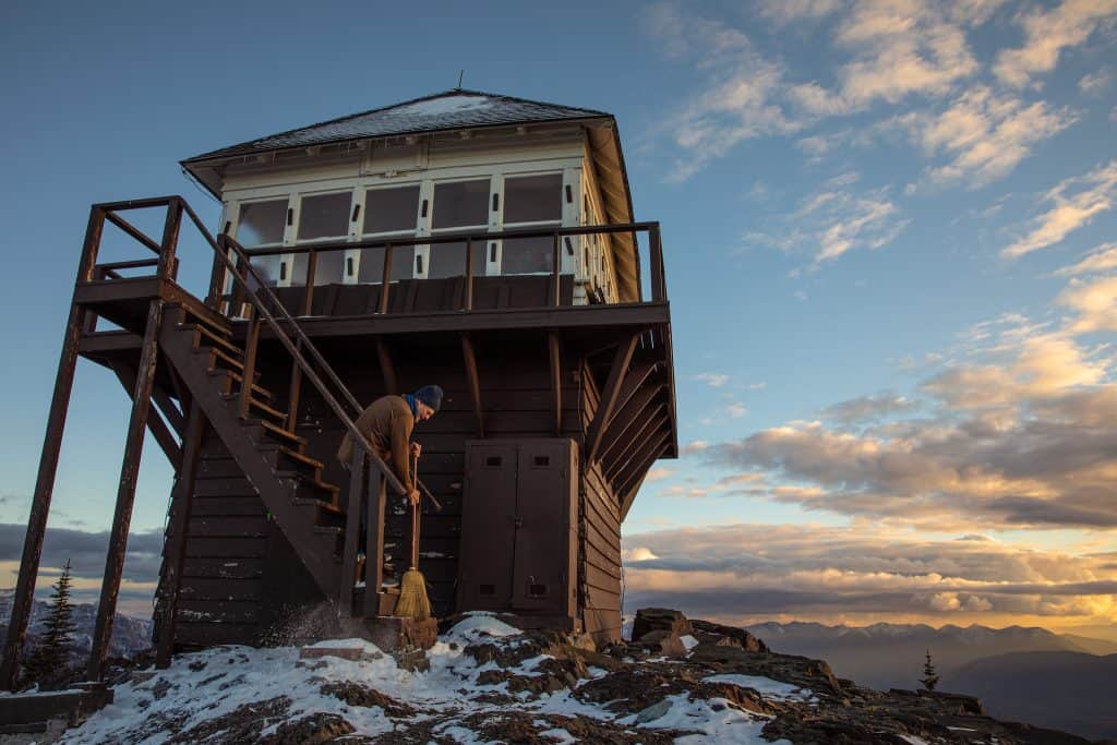 A person in a hat uses a broom to sweep snow from the bottom step of Mount Brown Lookout in Glacier National Park with mountains in the background.